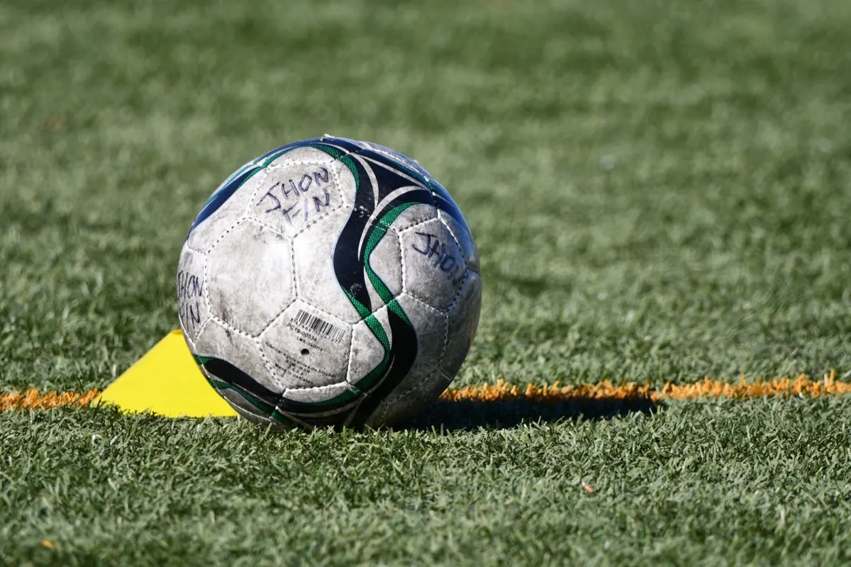 A worn match ball on a training pitch beside a yellow cone, with a child's name written on two of its panels in marker pen.