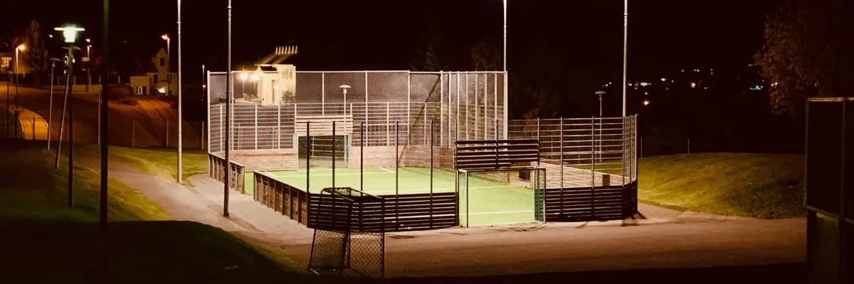 A small community pitch fenced and lit by floodlights on a dark evening.
